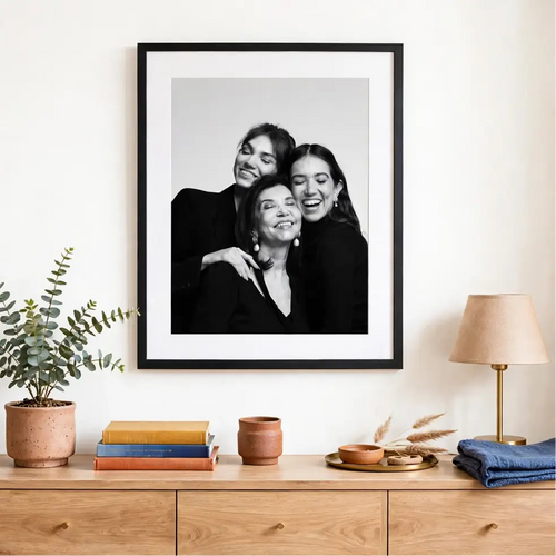 Un cuadro en blanco y negro enmarcado en negro, mostrando tres mujeres sonrientes de una familia, colocado sobre una consola con una lámpara en un espacio acogedor.